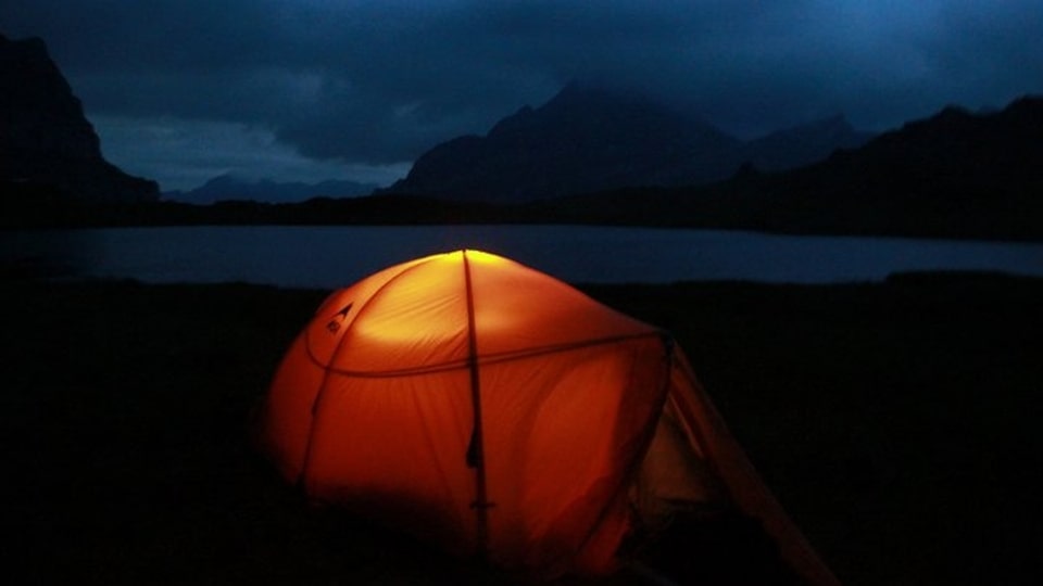 Ein Zelt steht nachts am Bergsee, von innen beleuchtet.