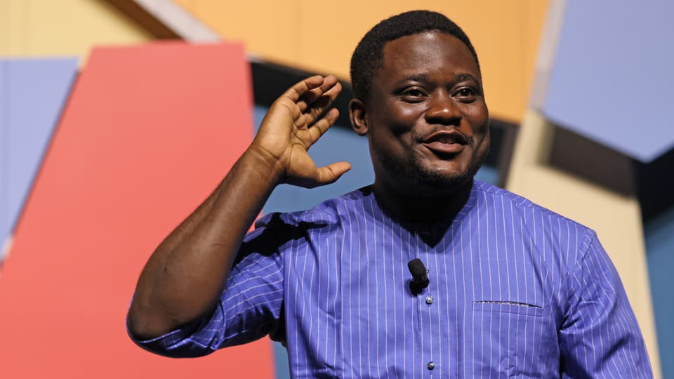 Bayo Akomolafe im blauen Hemd mit weissen Streifen trägt ein Mikrofon und hält seine Hand zum Ohr.