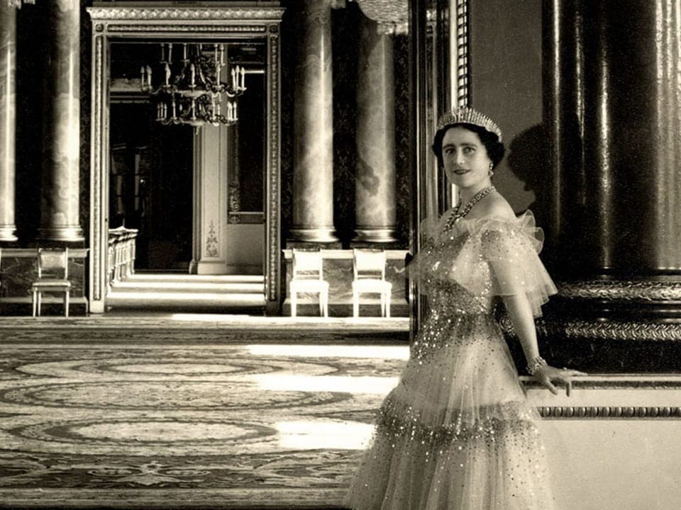 Queen Mum steht in einem weissen Kleid in einem Saal.