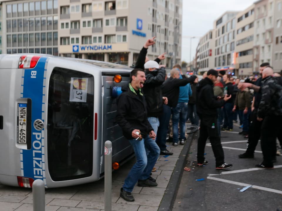 Hooligans posieren vor einem umgeworfenen Polizeiwagen
