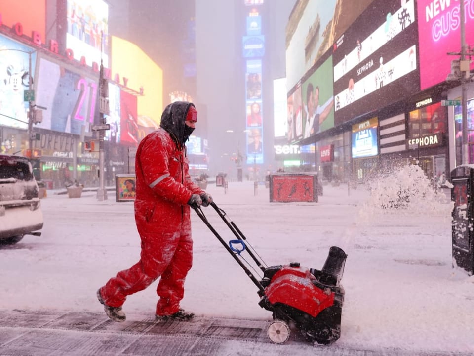 Ein Mann in orangenem Overall räumt mit einer Schneeschneuze den Schnee weg.