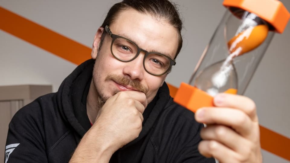junger Mann mit Brille und Schnurrbart hält grosses, orangenes Zeitglas, Kinn in Hand gestützt, schaut in Kamera.