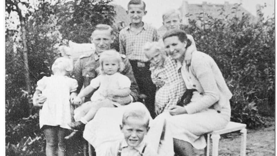 schwarzweiss Foto, rechts Frau im Rock, links Mann in Uniform, 6 kleine Kinder.