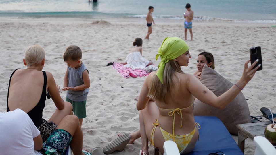 Eine Frau im Bikini mit hellgrünem Kopftuch macht ein Foto, im Hintergrund weitere Touristen, spielende Kinder am Meer