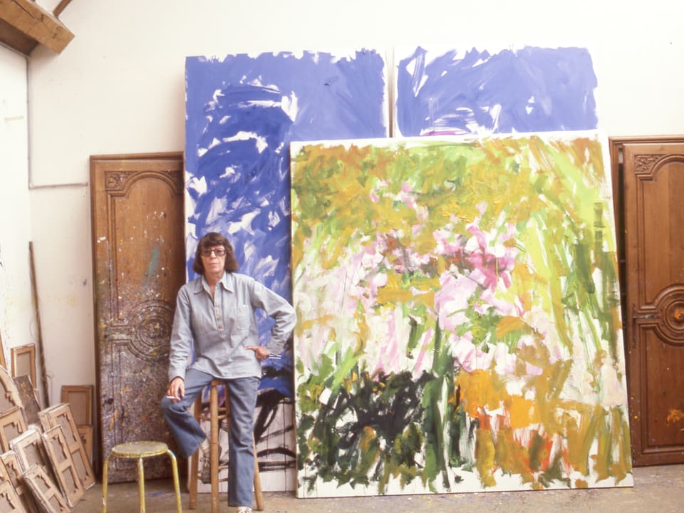 Eine Frau stitzt in einem Atelier vor zwei Gemälden. 