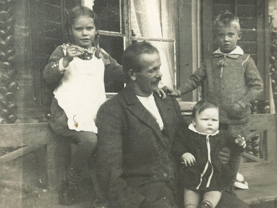 Drei kleine Kinder mit Vater vor Bauernhaus.