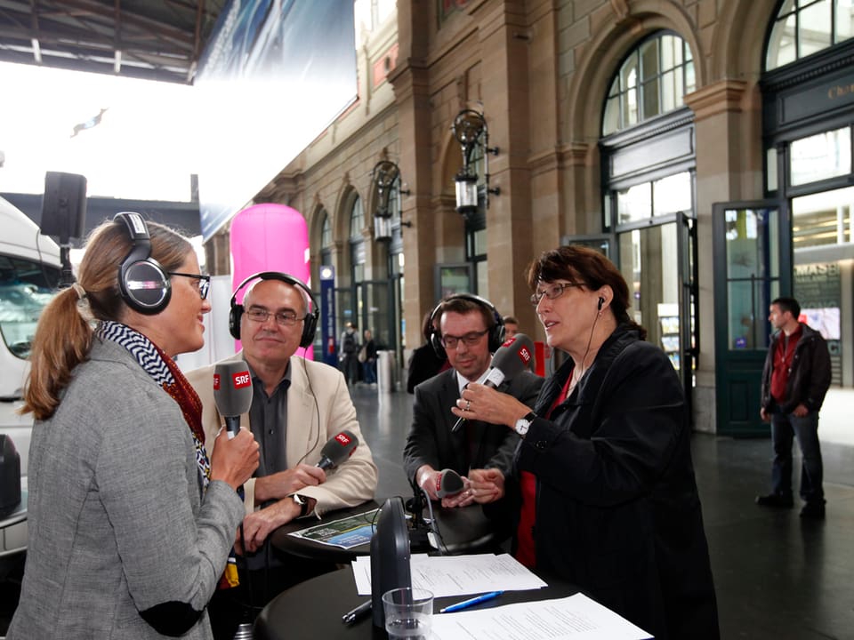 Zwei Frauen, zwei Männer um Stehtisch in der Halle des Zürcher Hauptbahnhofes (mit Mikrofonen)