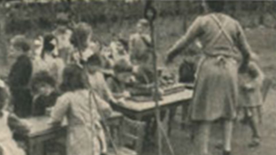Archivbild mit Kindern an einem Tisch, die von einer älteren Frau betreut werden.