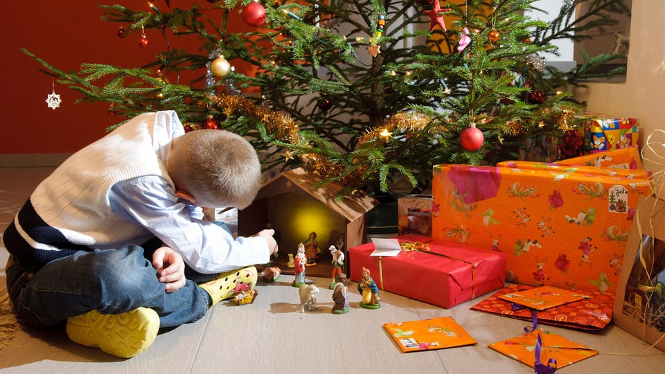 Ein Junge sitzt vor einem Weihnachtsbaum, unter dem mehrere Geschenke liegen.