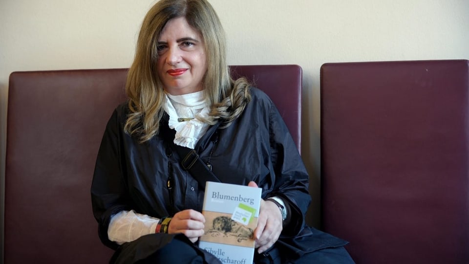 Lewitscharoff sitzt auf einem Stuhl mit ihrem Buch in der Hand.