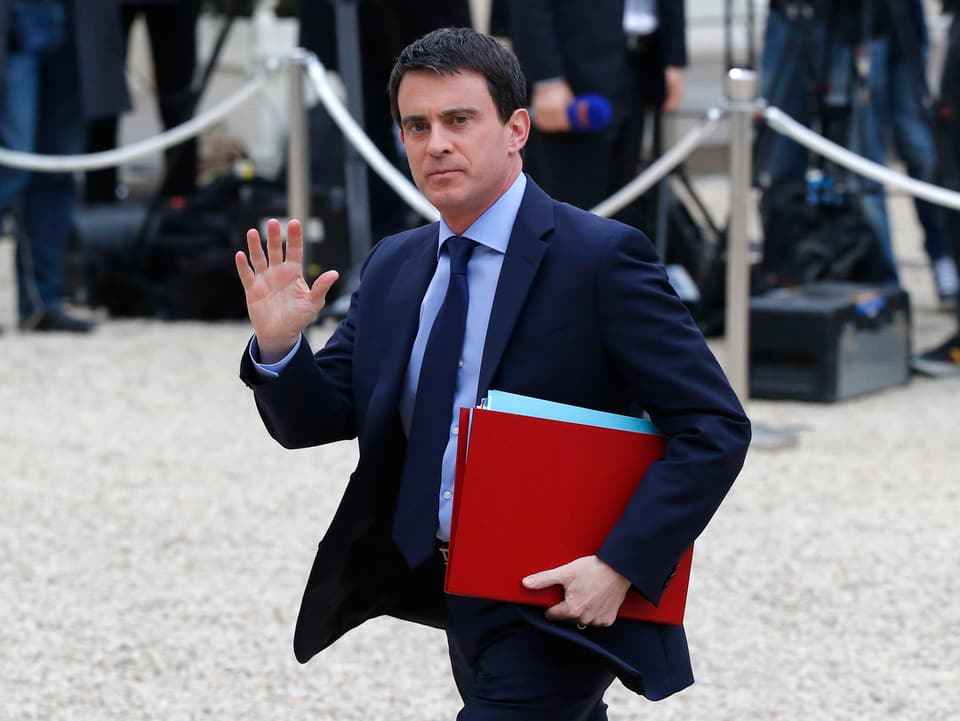Manuel Valls in einem Anzug und einer roten Mappe unter dem Arm. Mit der rechten Hand winkt er der Presse zu.