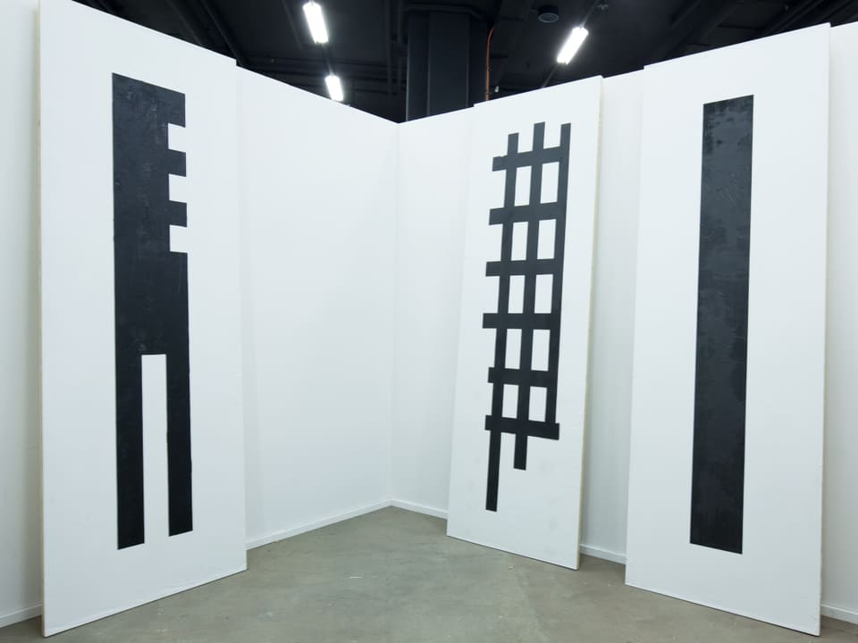Drei hohe, weisse Bretter mit geometrischer, schwarzer Bemalung lehnen an einer weissen Wand.