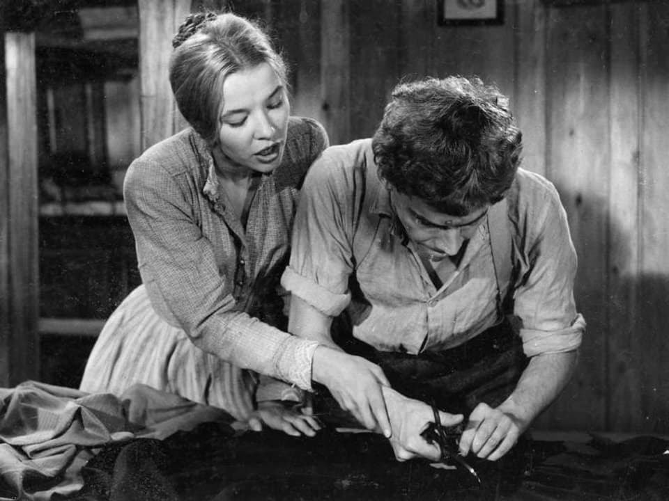 Szene aus Schwarzweiss-Film: Frau hilft Mann beim Stoffschneiden.