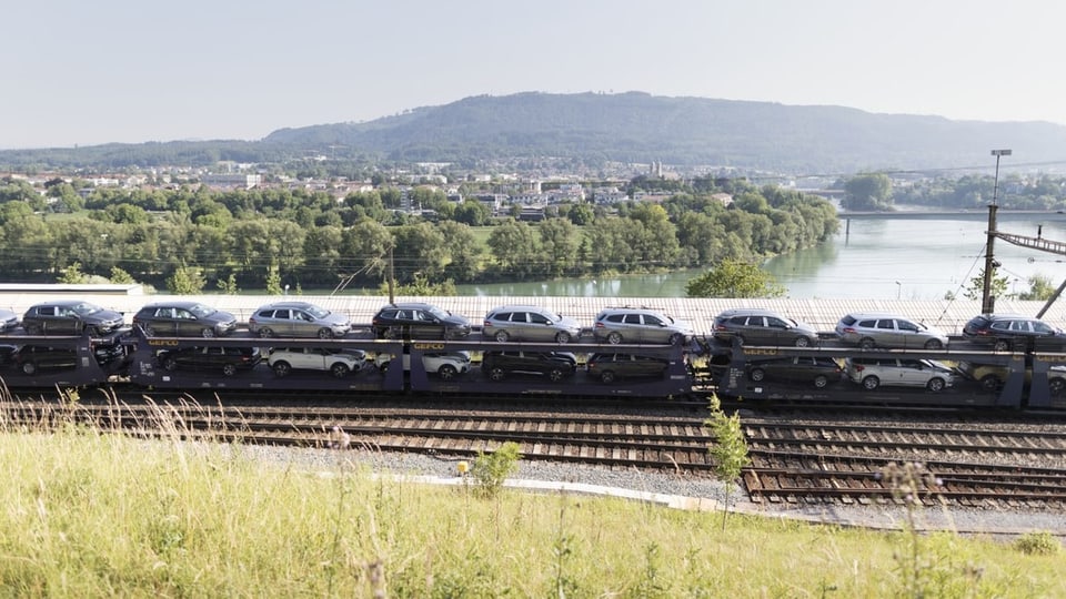 Ein Güterzug fährt an einem Fluss vorbei. Er hat Autos geladen.