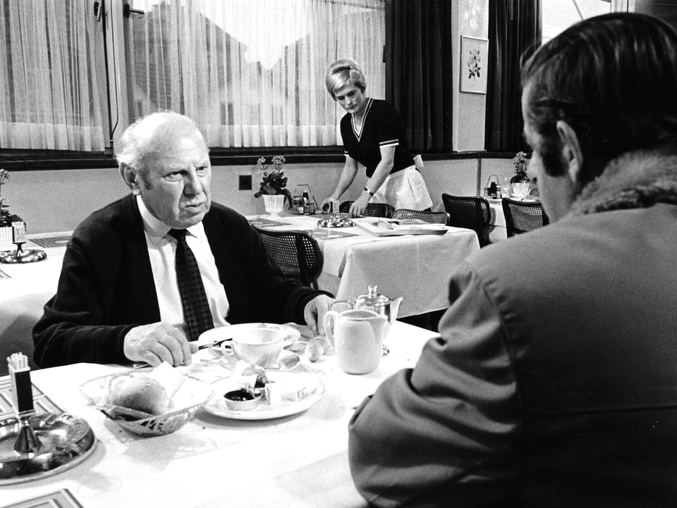 Ein alter Mann sitzt am Esstisch vor seinem Teller und blickt ernst auf den Mann, der ihm gegenüber sitzt.