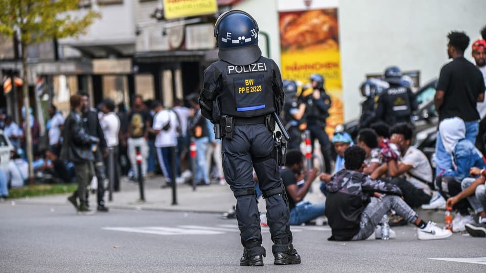 Ein Polizist steht mit dem Rücken zur Kamera. Vor ihm tummeln sich viele Menschen, daneben sitzen einige am Boden.