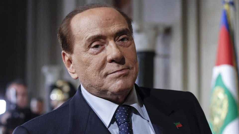 Silvio Berlusconi trägt einen Anzug mit Krawatte und schaut in die Kamera. 