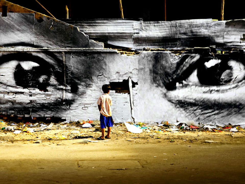 Junge steht vor einer Mauer mit einem Bildausschnitt eines Gesichtes (Augen und Nase) 