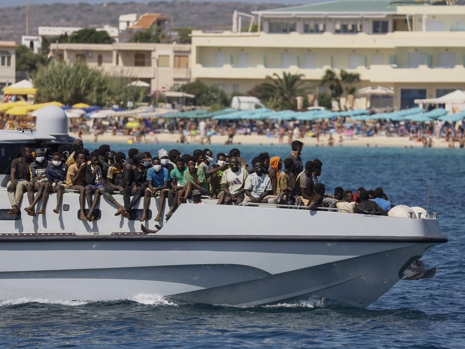 Ein Boot mit Flüchtlingen vor einem Strand mit Touristen. 