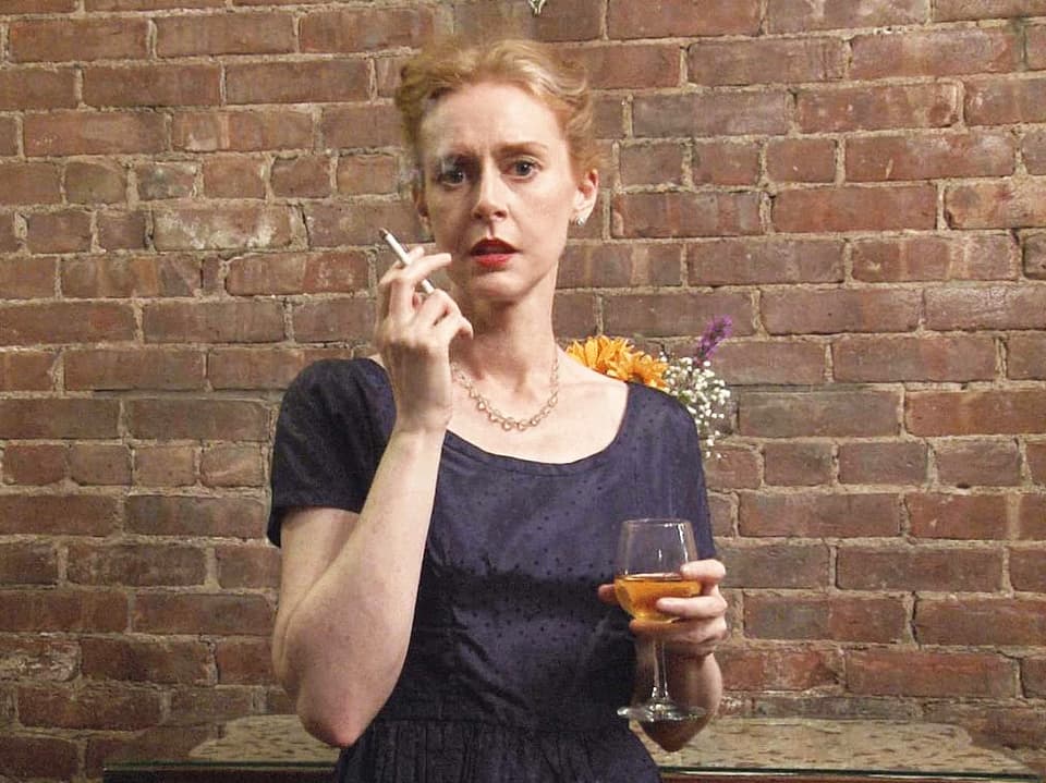 Eine Frau mit rotem, zusammengebundenem Haar. Sie trägt ein Weinglas und raucht.