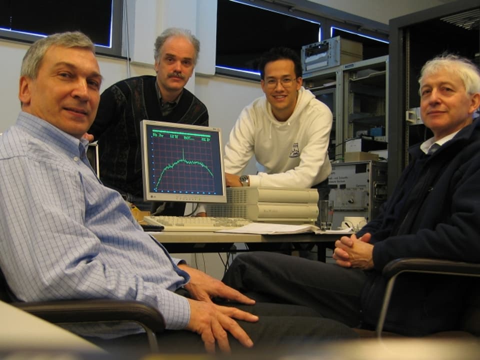 Vier Männer sitzen um einen Computer, auf dessen Bildschirm die grafische Darstellung eines Signals zu sehen ist.