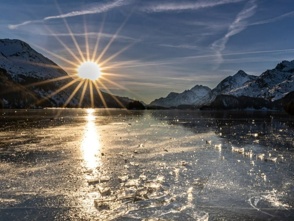 Silsersee/GR Blick über eine See mit Schwarzeis auf Berge, wo die Sonne gerade am klaren Himmel untergeht.