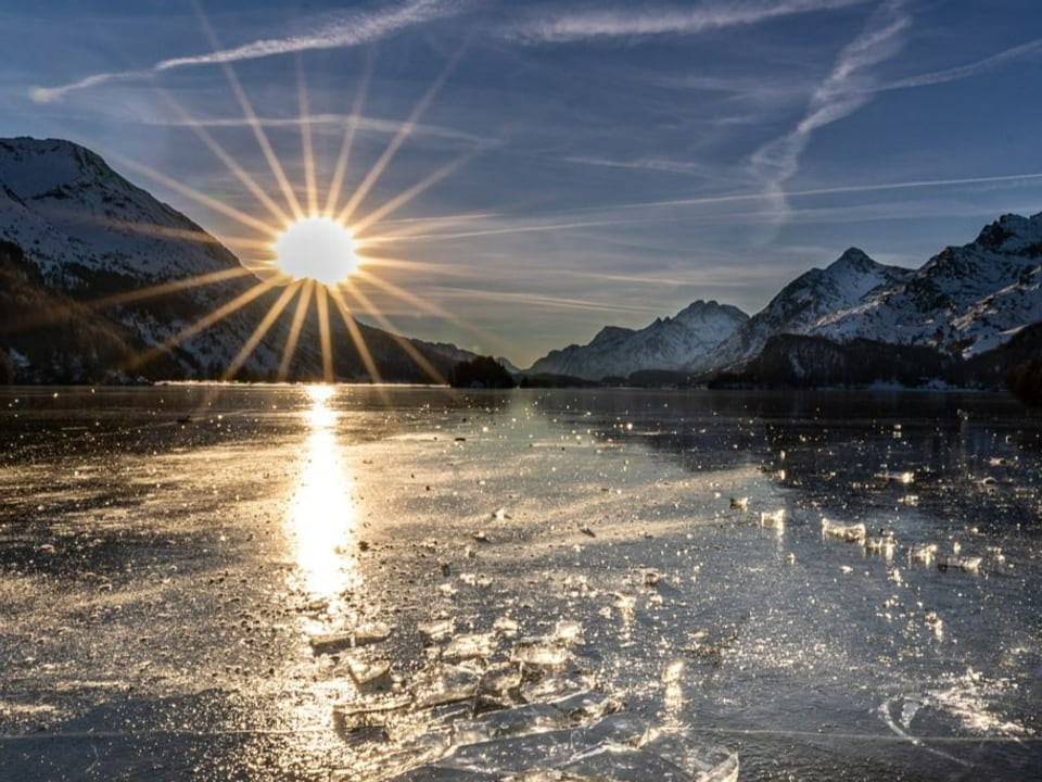 Blick über eine See mit Schwarzeis auf Berge, wo die Sonne gerade am klaren Himmel untergeht.