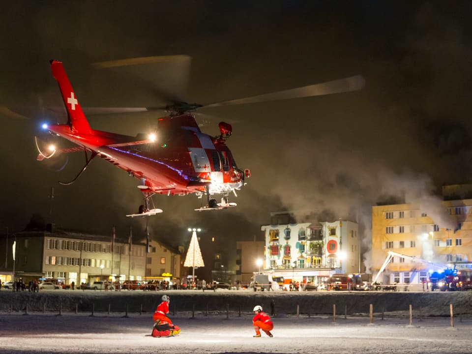 Rettungshelikopter landet auf gefrorenem Boden vor rauchendem Hotel