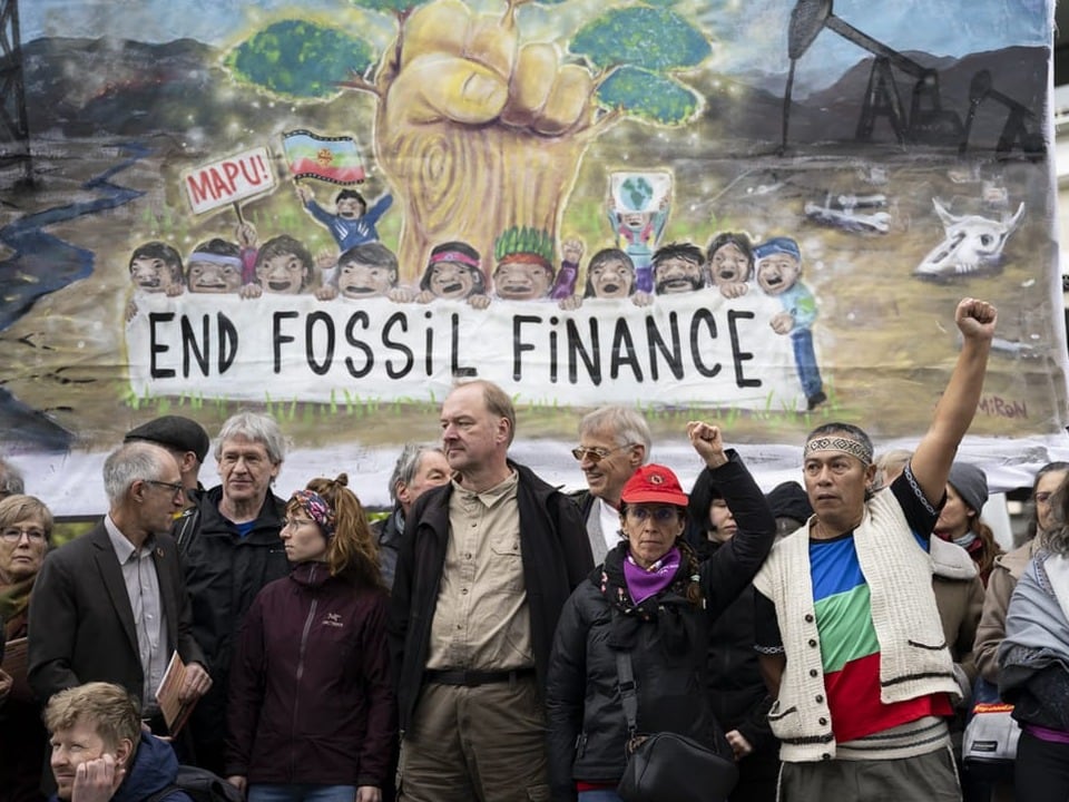 Demonstrierende halten ihre geballten Fäuste hoch, im Hintergrund ein Banner mit dem Titel «End Fossil Finance»