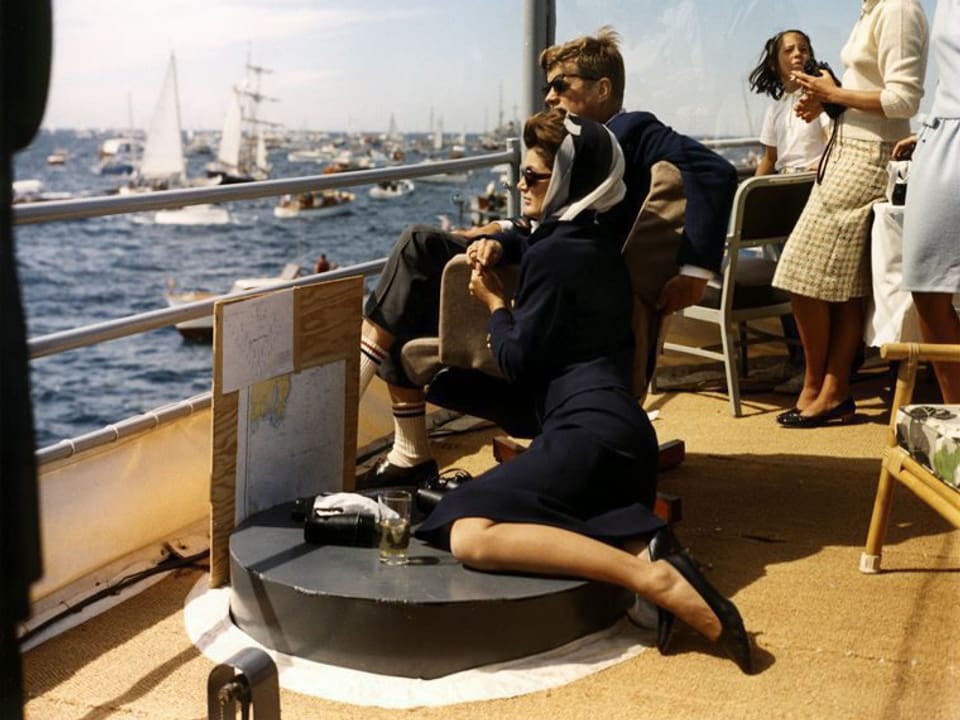 Ein Mann und eine Frau mit Sonnenbrille sitzen auf Deck eines Bootes und schauen einem Segelwettkampf zu.