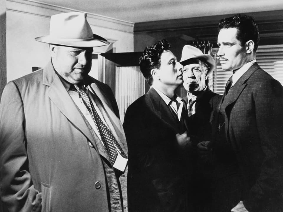 Schwarzweissbild, Filmszene. Welles mit hellem Mantel und Hut neben drei Männern in schwarzen Anzügen.