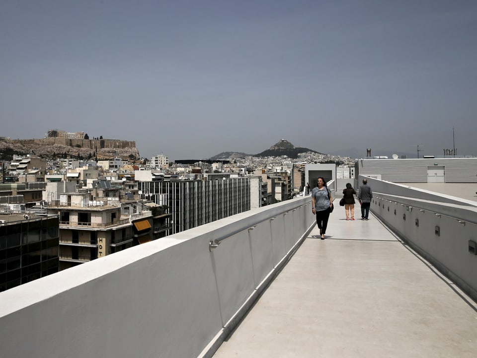 Leute laufen über die Terasse eines Museums in Athen. Im Hintergrund sieht man die Stadt.
