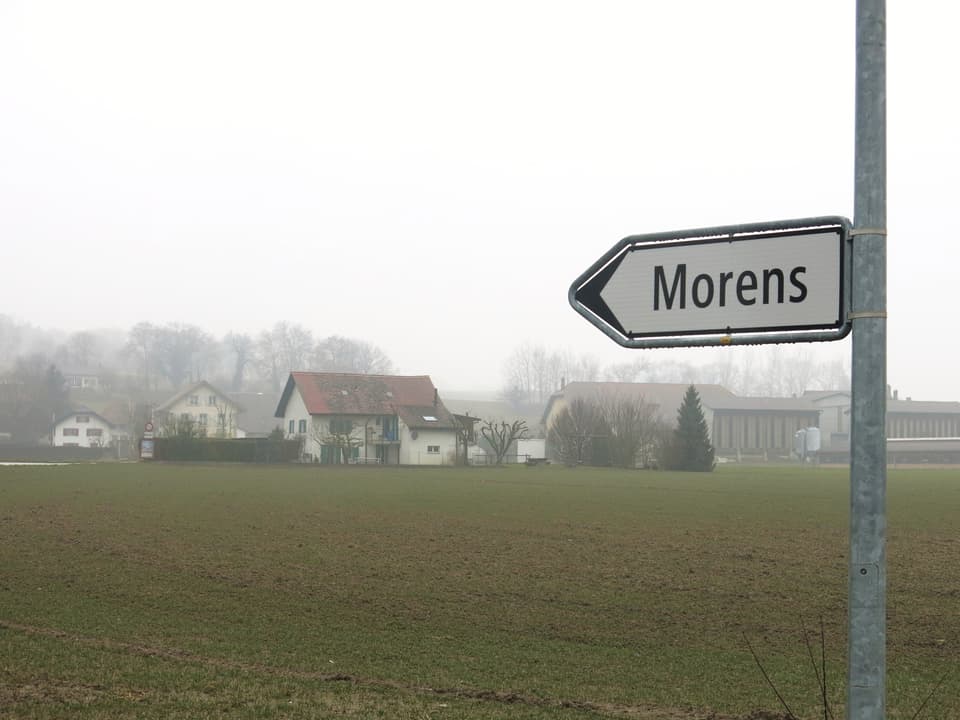 Im Vordergrund ein Wegweiser nach Morens, im Hintergrund ein Wohnhaus in einem Feld.
