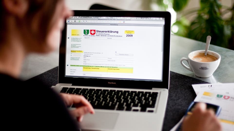 Eine Person sitzt an einem Tisch und füllt am Computer eine Steuererklärung aus. 