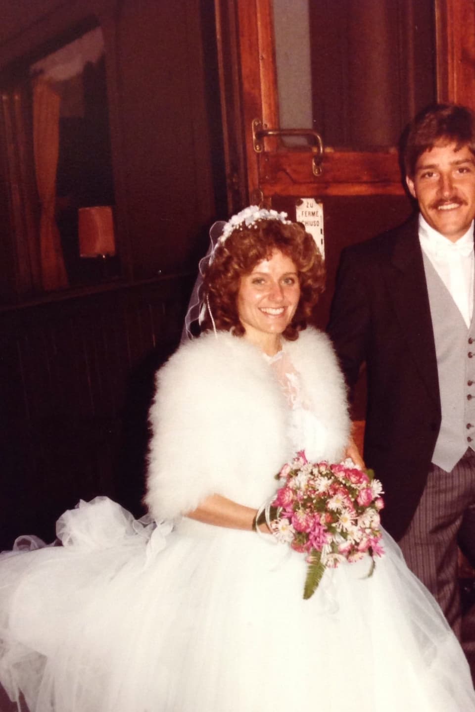Ein lächelndes Brautpaar, sie im Kleid mit einer Schleppe aus Tüll, er im Frack, steht am Aufstieg eines alten Zugwaggons und lächelt in die Kamera.