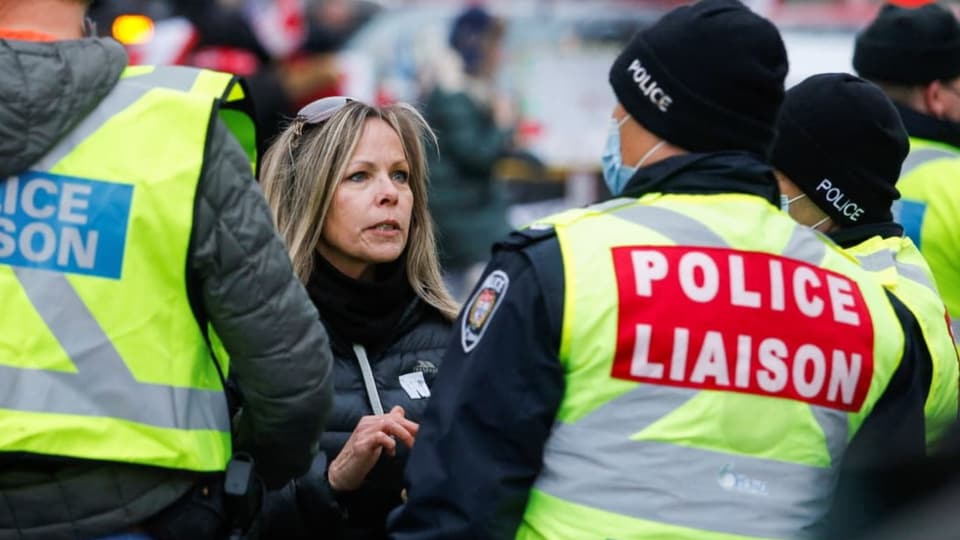 Tamara Lich, lange Haare, Sonnenbrille im Haar, zwischen zwei Polizisten mit Leuchtwesten.