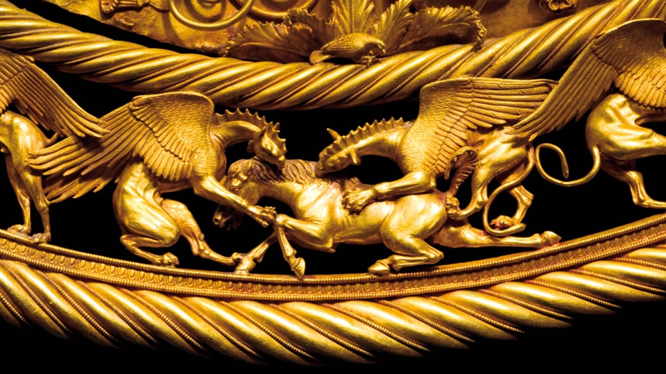 Eine goldene Reliquie, die geflügelte Drachenwesen im Kampf mit einem Pferd darstellt.