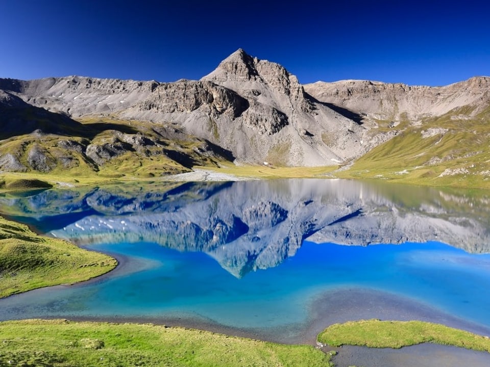 Bergsee mit Gipfel, der sich darin spiegelt. Der Himmel ist stahlblau. 