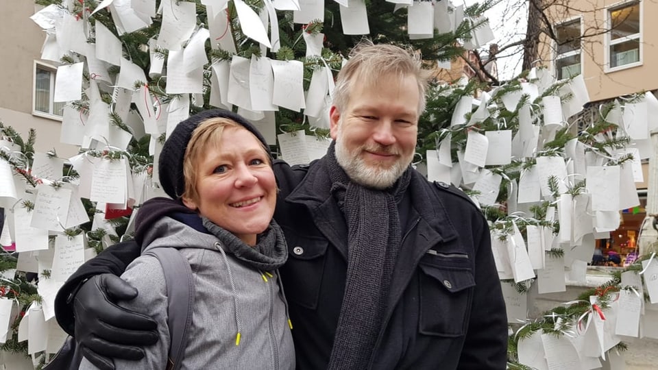 Foto Rechts Mann, hält Frau, links, im Arm. Im Hintergrund ein Baum, vollbehängt mit weissen Zettelchen.