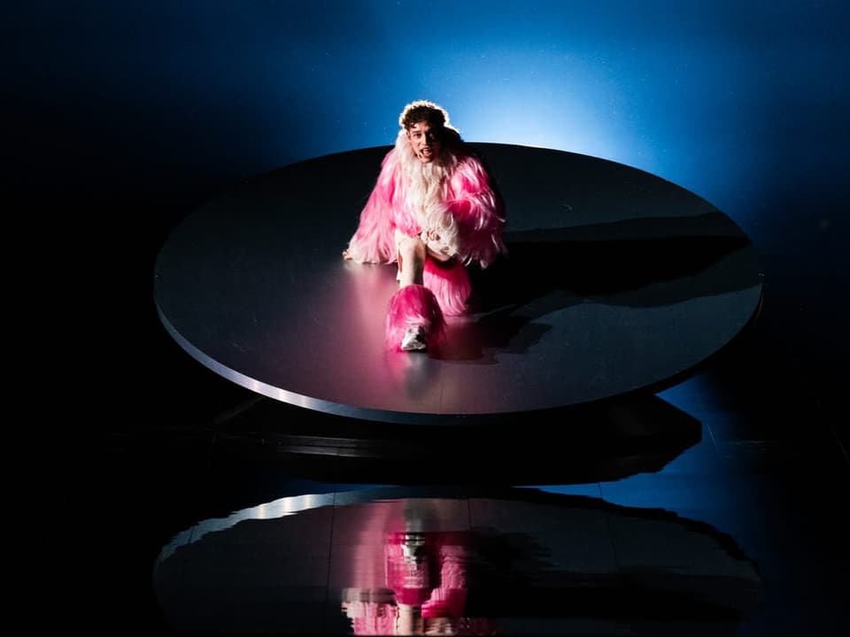 Person in rosa Federkostüm sitzt auf einer runden Bühne unter blauem Licht mit Spiegelung.