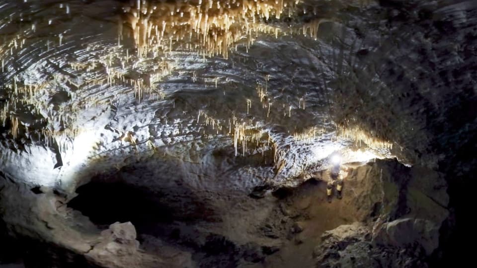 Eine Höhle, beleuchtet von der Stirnlampe einer Person, die aus einem Seitengang kommt. Von der Decke hängen Stalaktiten