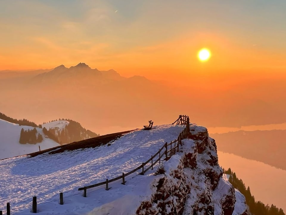 Blick von einem Berg mit Schnee ins Mittelland, wo die Sonne mit farbigem Himmel untergeht. 