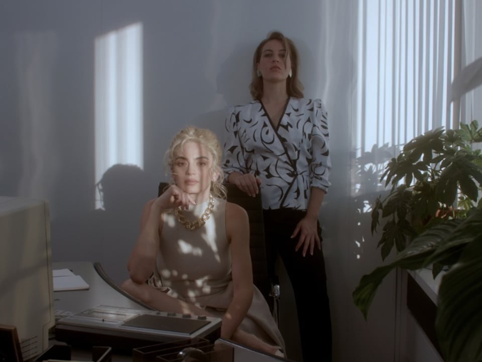 zwei junge Frauen sitzen und stehen hinter Schreibtisch, schauen lasziv in die Kamera, eine hat den Ellbogen aufgestützt
