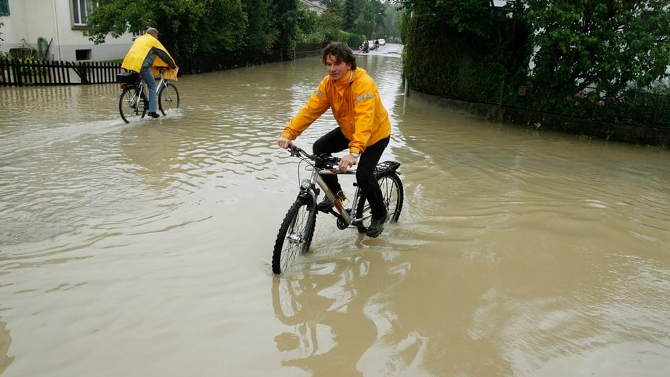 Eine überschwemmte Quartierstrasse, auf der zwei Fahrräder durch die Wassermassen pflügen. Das Wasser steht den Velofahrern dabei fast bis zu den Pedalen.