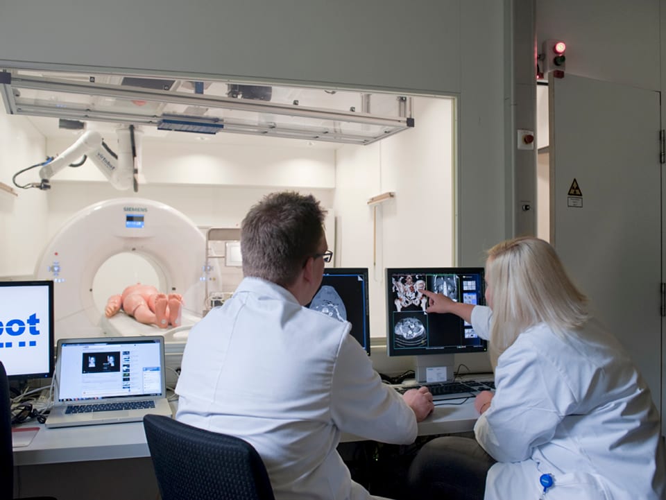 Zwei Personen in Laborkleidung betrachten den Monitior eines CT im Nebenraum