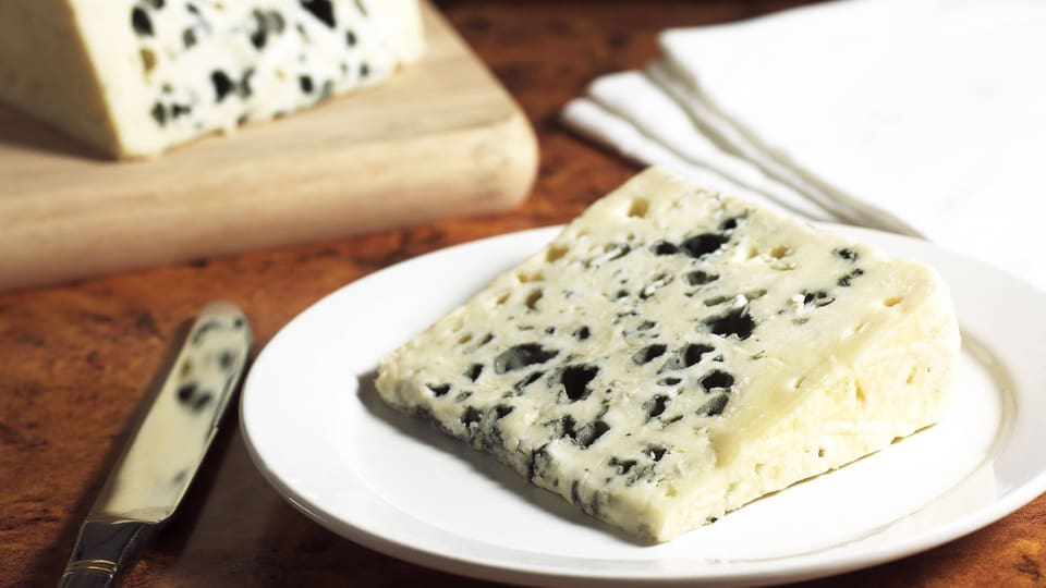 Heller Käse mit dunkelgrünen Schimmelstücken, liegt auf Brett, daneben Messer.