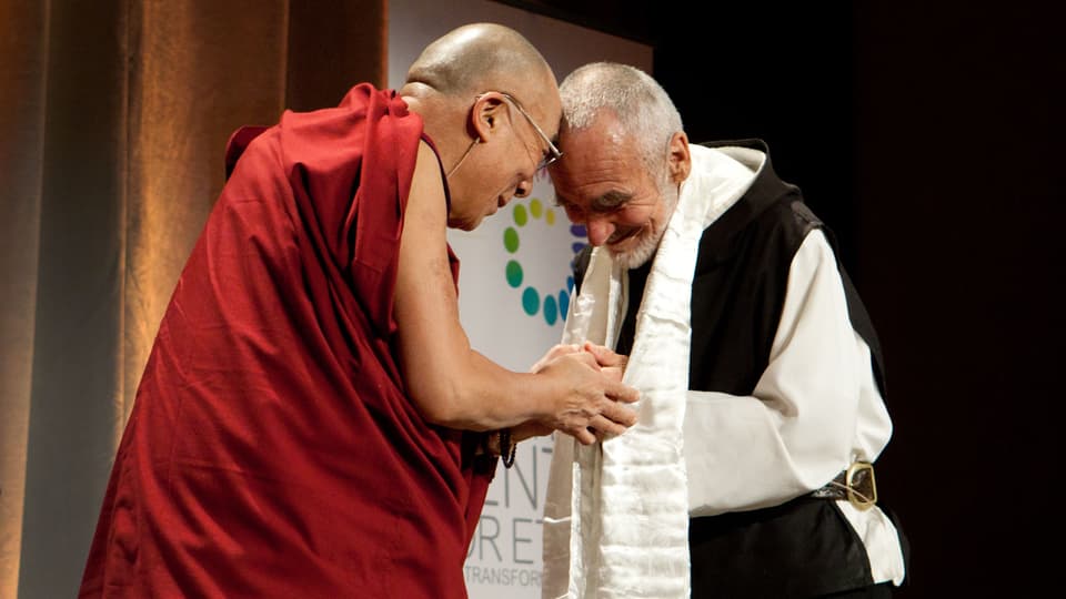 Dalai Lama (links, in rot) und David Steindl-Rast begrüssen sich, halten Hände und Kopf zusammen, lächeln.