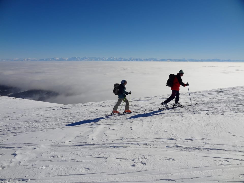 Zwei Skitourengänger erklimmen den Chasseral. Das Mittelland ist geflutet mit Hochnebel. Am Horizont sind die Alpen zum Greifen nahe. Darüber wolkenloser Himmel.