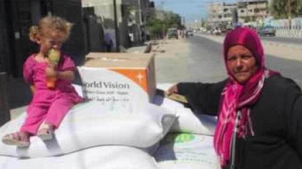 Ein Mädchen sitzt auf weissen Lebensmittelsäcken vor einer Schachtel mit dem World-Vision-Logo, daneben steht eine Frau mit Kopftuch.
