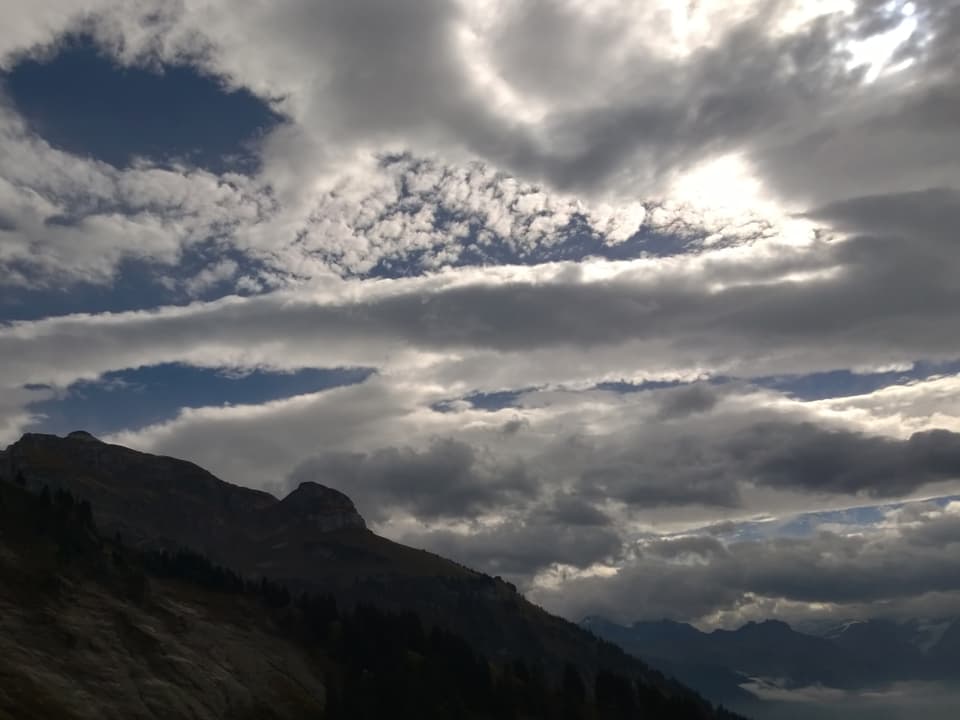 Das Bild zeigt in der unteren Hälfte ein Gebirgszug, der Fronalpstock im Glarnerland. In der oberen Hälfte wird der blaue Himmel von bänderartigen Wolkenfeldern überzogen. Die Sonne versteckt sich hinter einem Wolkenband.